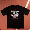A black short-sleeve athletic T-shirt laid flat on a red running track, featuring a large front graphic with white and red text reading 'HYBRID ATHLETE' and card icons. Parts of a person's legs and shoes are visible around the track.