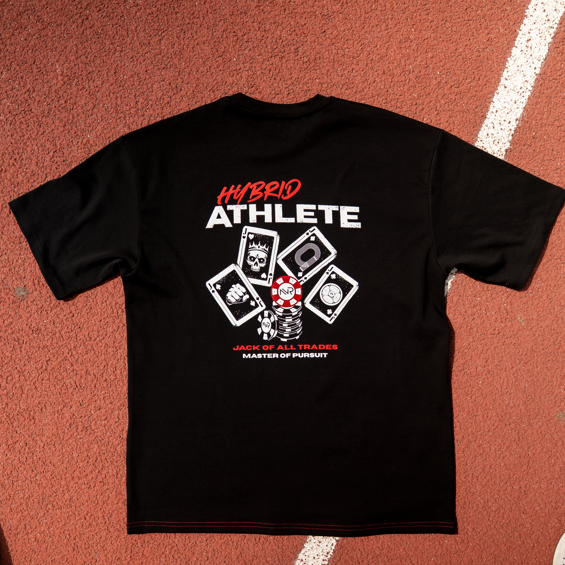 A black short-sleeve athletic T-shirt laid flat on a red running track, featuring a large front graphic with white and red text reading 'HYBRID ATHLETE' and card icons. Parts of a person's legs and shoes are visible around the track.