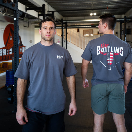 Two men in a gym setting, one facing the camera and the other with a nvr rest shirt on the back.