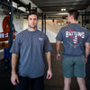 Two men in a gym setting, one facing the camera and the other with a nvr rest shirt on the back.